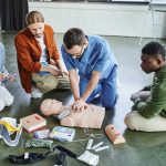 Medical instructor in blue clothing demonstrates chest compressions for CPR in front of three learners.