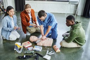 Medical instructor in blue clothing demonstrates chest compressions for CPR in front of three learners.