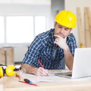 A construction projects manager sits down at a table, with tools and PPE on it, to work on his NVQ portfolio at his laptop. He is wearing a yellow hard hat.