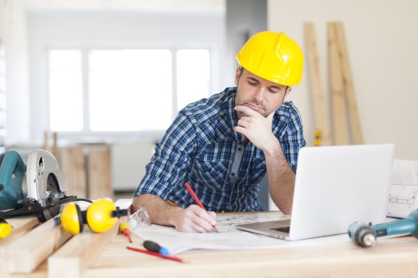 A construction projects manager sits down at a table, with tools and PPE on it, to work on his NVQ portfolio at his laptop. He is wearing a yellow hard hat.