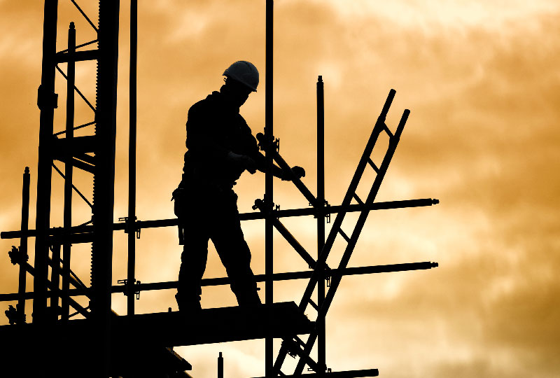 Silhouette of a builder erecting scaffolding against an orange sky.