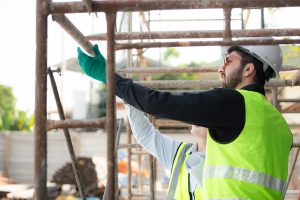 A joint sealant worker in PPE erecting scaffolding with another worker helping.