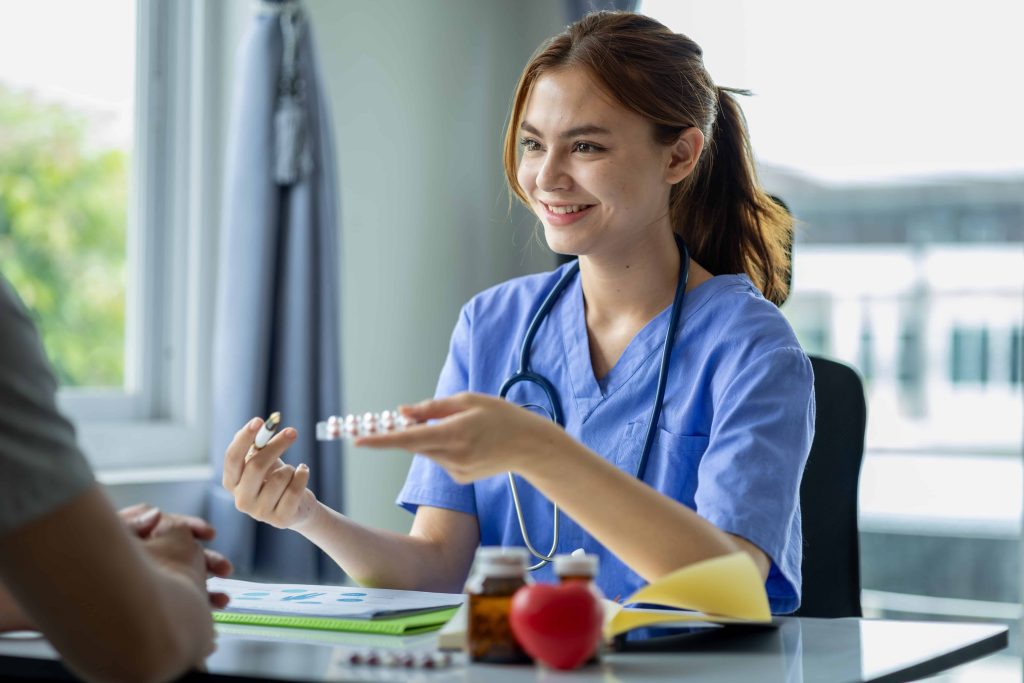 Healthcare professional wearing blue scrubs and a stethoscope, holding a blister pack of pills while discussing with another person. The desk has medication bottles, an open notebook, and a red heart-shaped object, with natural light coming through a window.