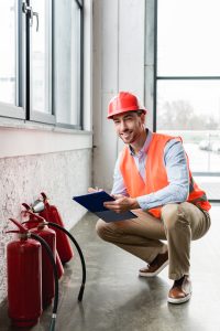 A person wearing an orange safety vest and a hard hat squats down while holding a blue clipboard, inspecting three red fire extinguishers mounted on the wall. The scene is indoors, near large windows with natural light coming in.