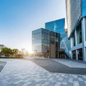 Modern glass office buildings with a wide open plaza in front, photographed at sunrise with trees in the background.