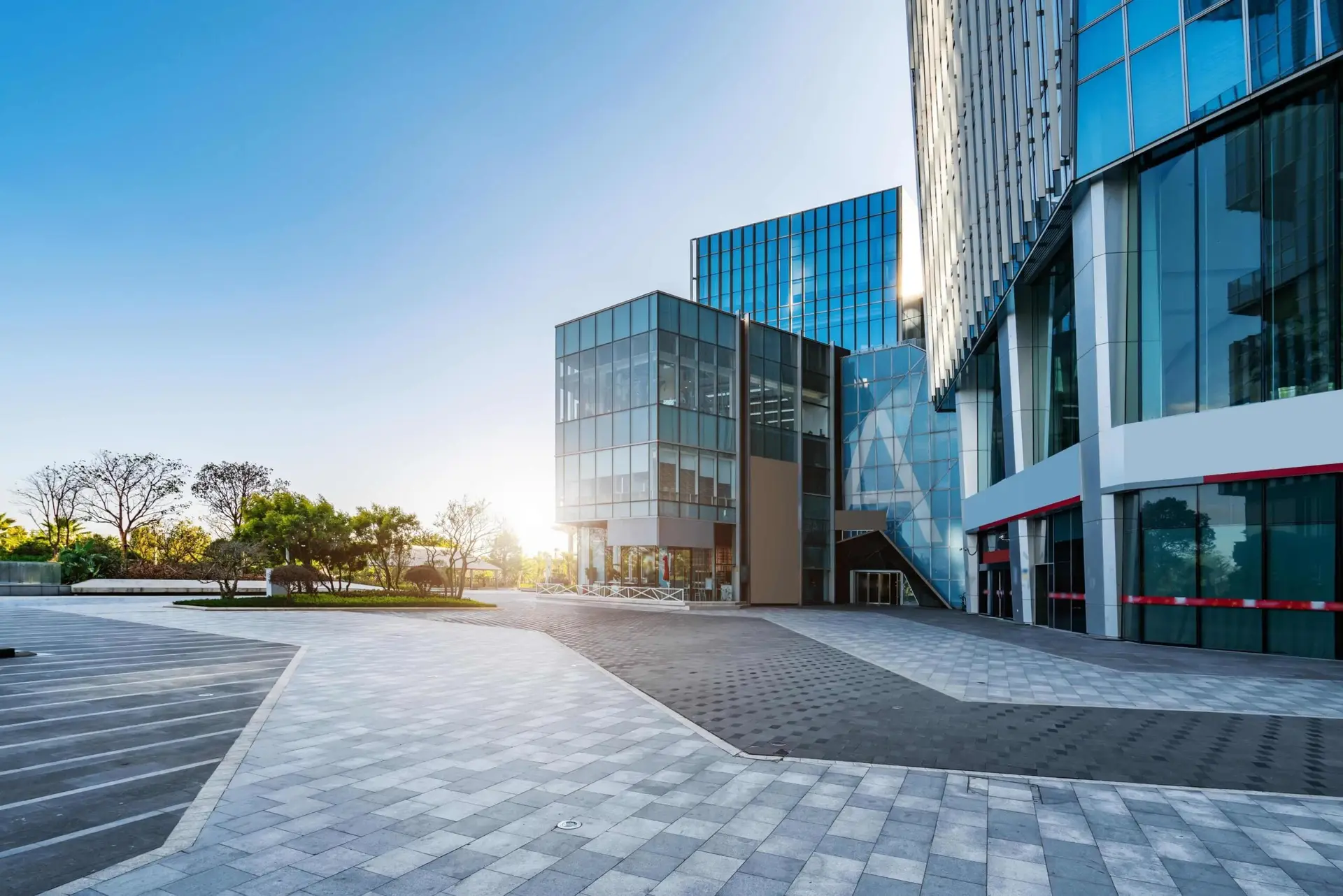 Modern glass office buildings with a wide open plaza in front, photographed at sunrise with trees in the background.