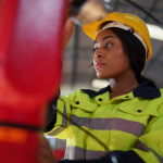 A person wearing a yellow hard hat and a high-visibility jacket operates machinery in an industrial setting, ensuring fire safety protocols are followed. The background shows the interior of a large building with metal beams and lighting fixtures.