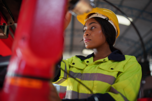 A person wearing a yellow hard hat and a high-visibility jacket operates machinery in an industrial setting, ensuring fire safety protocols are followed. The background shows the interior of a large building with metal beams and lighting fixtures.
