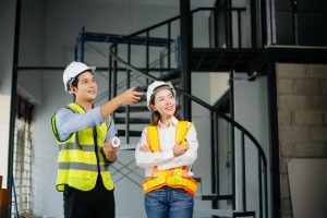 Two construction workers in safety helmets and reflective vests inside a building under construction, with one pointing while holding blueprints and the other observing; scaffolding and metal stairs in the background.