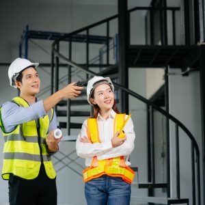 Two construction workers in safety helmets and reflective vests inside a building under construction, with one pointing while holding blueprints and the other observing; scaffolding and metal stairs in the background.