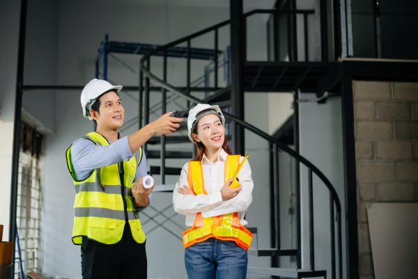 Two construction workers in safety helmets and reflective vests inside a building under construction, with one pointing while holding blueprints and the other observing; scaffolding and metal stairs in the background.