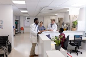 Medical professionals in a hospital setting discuss patient care at a reception counter, with a wheelchair and medical equipment visible in the background.