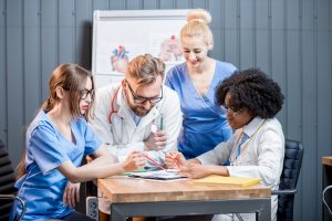 The image shows a group of four medical professionals, including doctors and nurses, gathered around a table in a discussion. They are looking at documents and charts on the table. In the background, there is a whiteboard with anatomical diagrams of the human heart and brain.