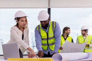 A group of construction professionals wearing safety helmets and reflective vests are gathered around a table with blueprints and a laptop, discussing project plans in a modern office setting.