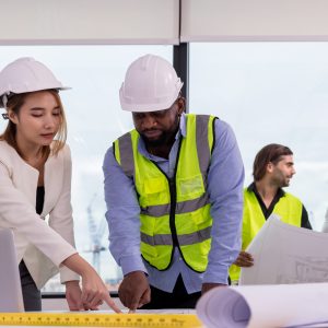 A group of construction professionals wearing safety helmets and reflective vests are gathered around a table with blueprints and a laptop, discussing project plans in a modern office setting.