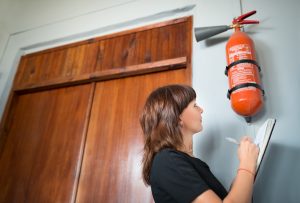 Person inspecting a red fire extinguisher mounted on a wall, holding a clipboard