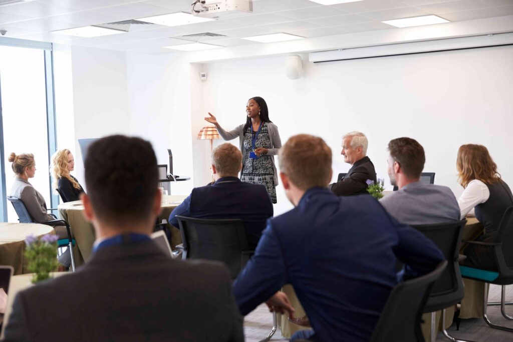 A person standing at the front of a bright, modern conference room giving a presentation to a group seated around tables. The presenter is gesturing with one hand while speaking, and attendees are facing forward, listening attentively. The room features white walls, a ceiling-mounted projector, and large windows allowing natural light.