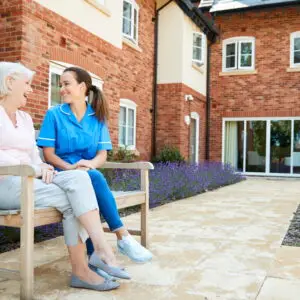 An elderly woman sitting on an outdoor bench with a care worker in front of a modern residential care home, surrounded by landscaped gardens and lavender plants.