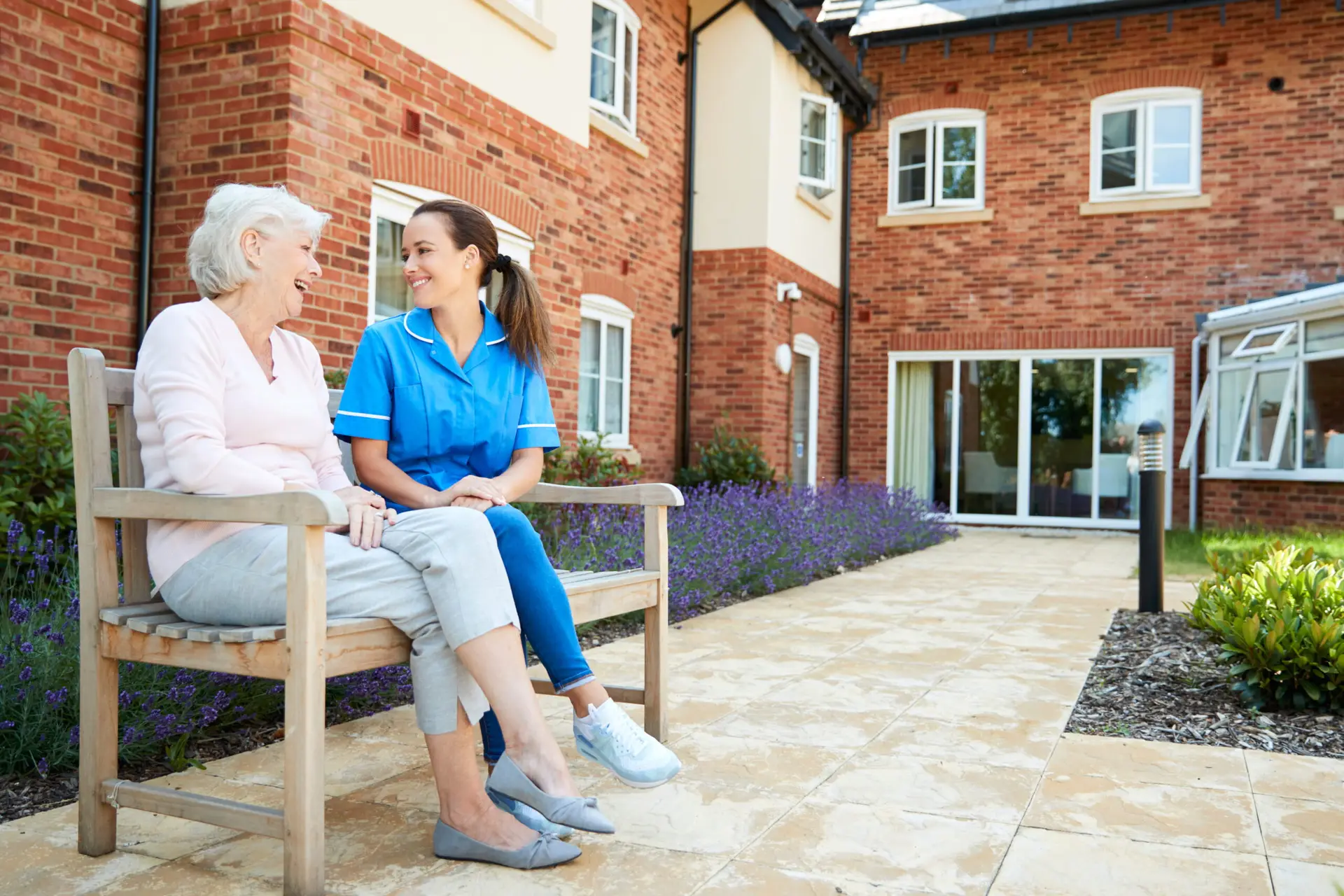 An elderly woman sitting on an outdoor bench with a care worker in front of a modern residential care home, surrounded by landscaped gardens and lavender plants.