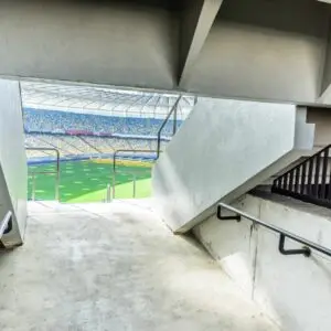 View from a stadium concourse looking out toward the pitch through a concrete exit tunnel, with handrails on both sides and a brightly lit football field visible in the distance.