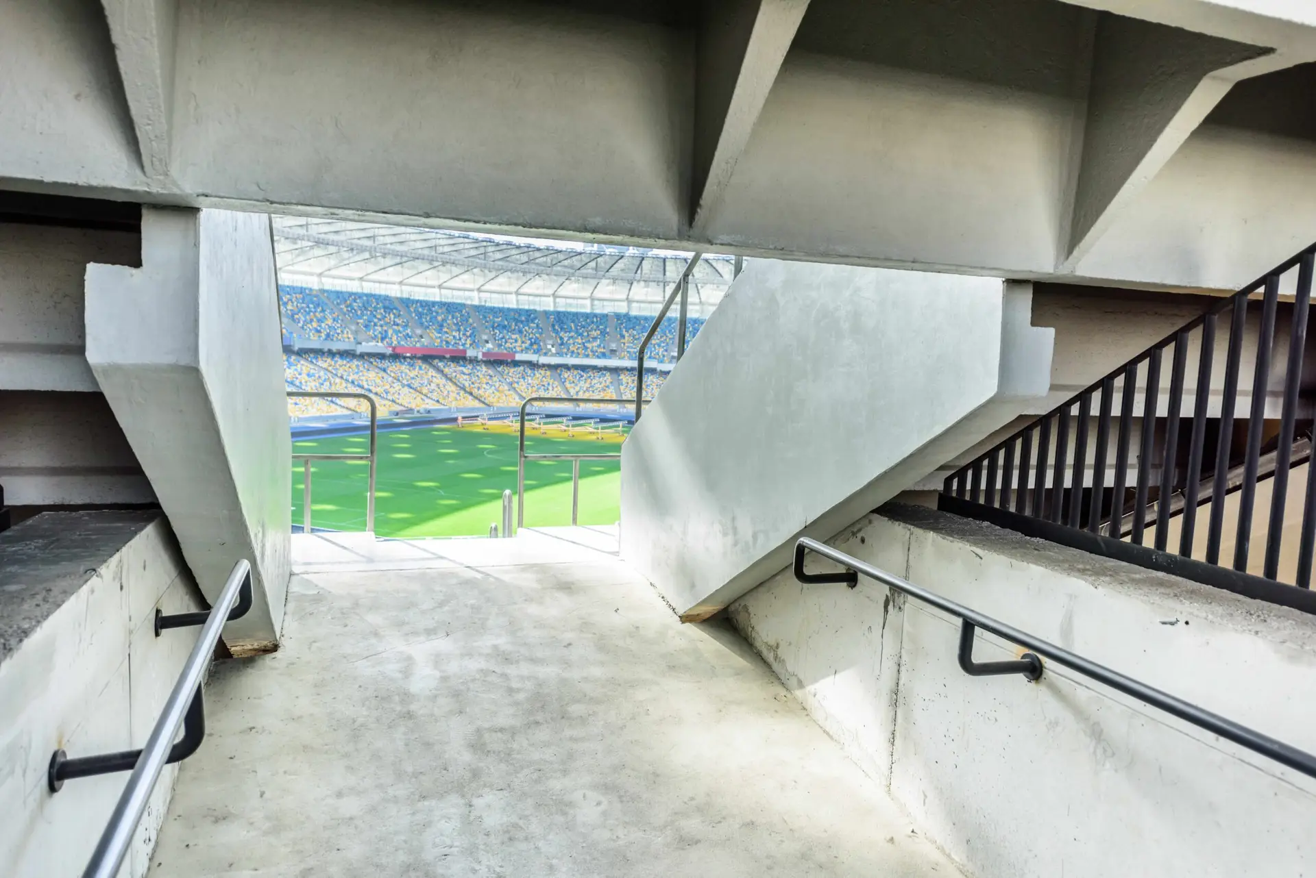 View from a stadium concourse looking out toward the pitch through a concrete exit tunnel, with handrails on both sides and a brightly lit football field visible in the distance.