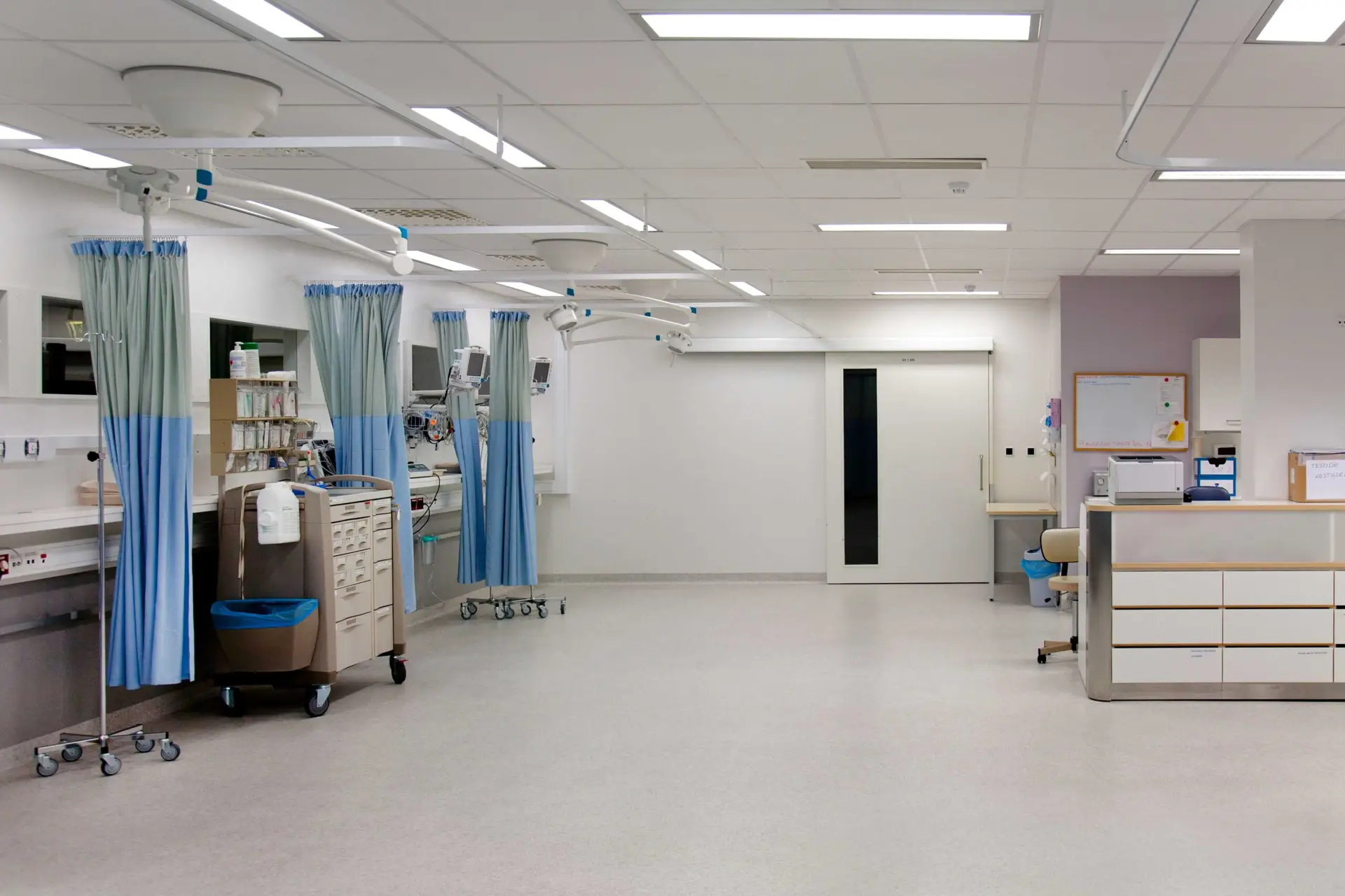 A clean, modern hospital ward with medical equipment, curtained treatment bays, storage units, and bright overhead lighting, designed for patient care and monitoring.