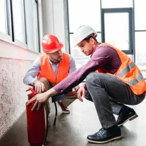 Two workers wearing safety helmets and high‑visibility vests kneeling indoors while inspecting a fire extinguisher near a large window.