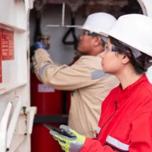 Two workers wearing protective helmets and safety gear inspecting fire equipment, with one person checking a fire extinguisher and the other holding a tablet for recording information.