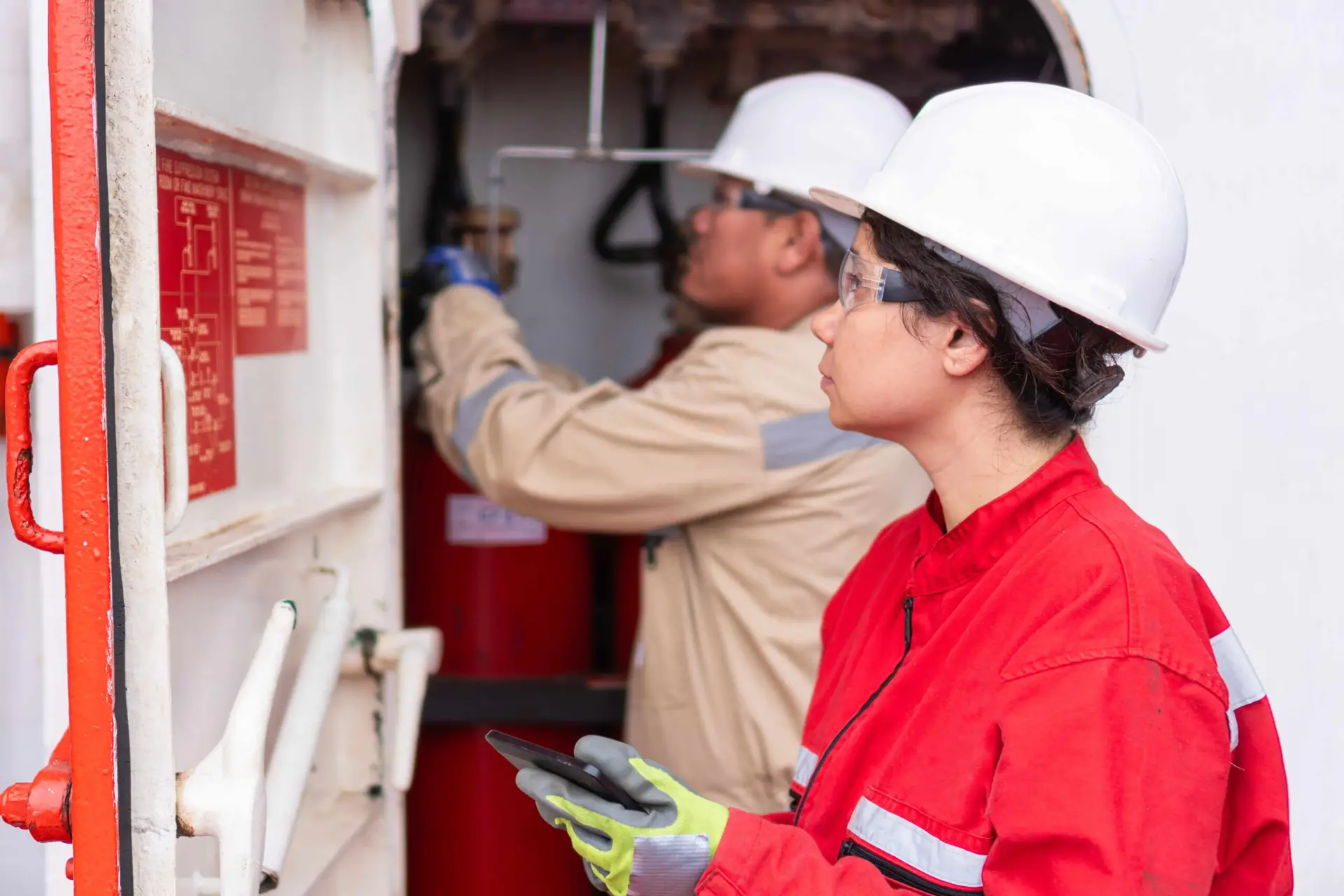 Two workers wearing protective helmets and safety gear inspecting fire equipment, with one person checking a fire extinguisher and the other holding a tablet for recording information.