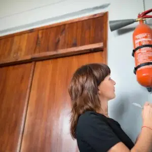 A person conducting a fire extinguisher inspection, writing notes on a clipboard while observing a wall‑mounted fire extinguisher.
