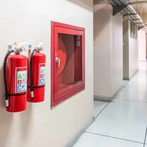 Two wall‑mounted ABC fire extinguishers and a red fire hose cabinet in a bright hallway of a commercial building.