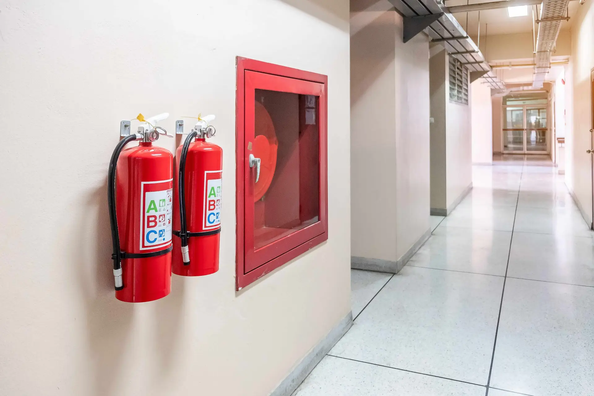 Two wall‑mounted ABC fire extinguishers and a red fire hose cabinet in a bright hallway of a commercial building.