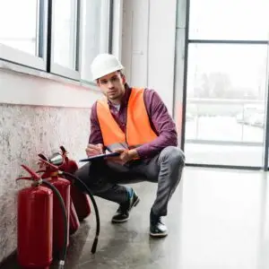 A safety inspector wearing a hard hat and high‑visibility vest kneeling to examine and record details of fire extinguishers using a clipboard inside a bright commercial building.