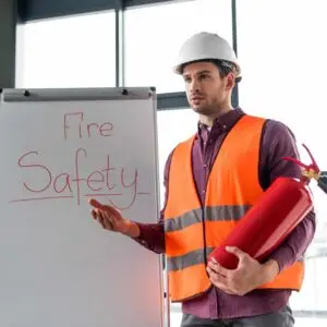 A fire safety instructor wearing a hard hat and high‑visibility vest holding a fire extinguisher while presenting beside a whiteboard labelled “Fire Safety.”