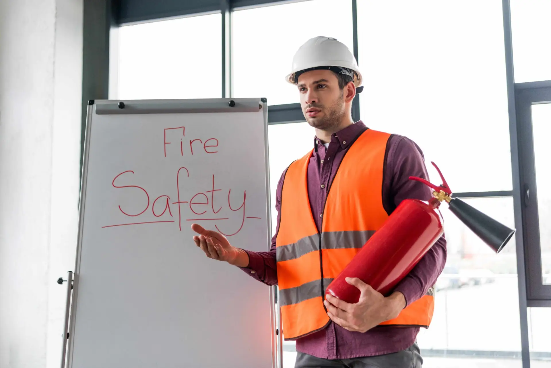 A fire safety instructor wearing a hard hat and high‑visibility vest holding a fire extinguisher while presenting beside a whiteboard labelled “Fire Safety.”