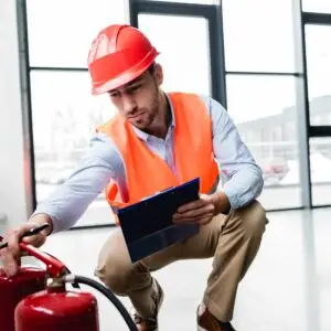 A safety inspector wearing a hard hat and high‑visibility vest kneeling to check a fire extinguisher while recording information on a clipboard inside a commercial building.