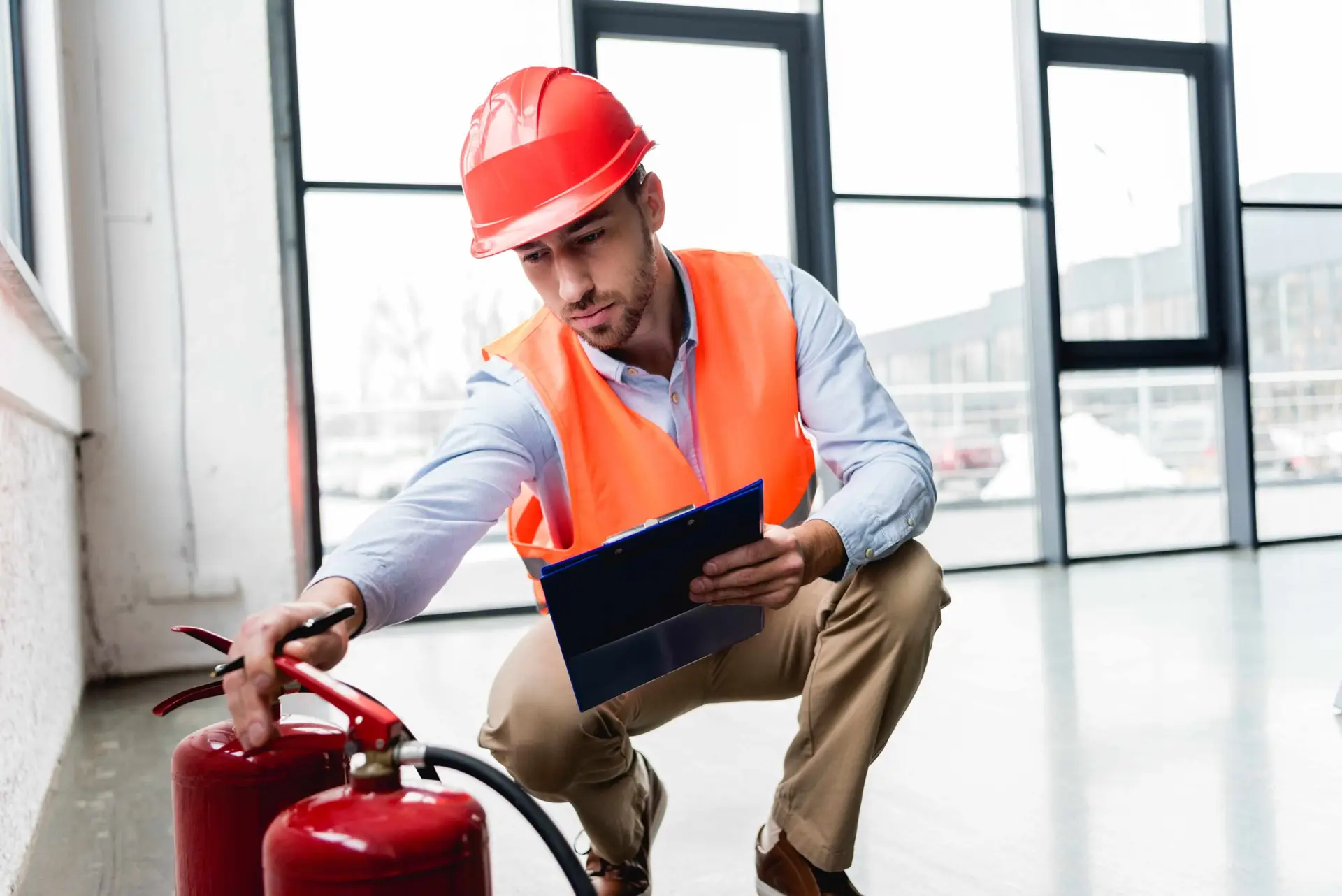 A safety inspector wearing a hard hat and high‑visibility vest kneeling to check a fire extinguisher while recording information on a clipboard inside a commercial building.