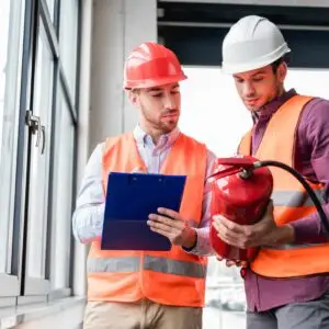 Two safety inspectors wearing hard hats and high‑visibility vests reviewing a clipboard together while examining a fire extinguisher inside a commercial building.