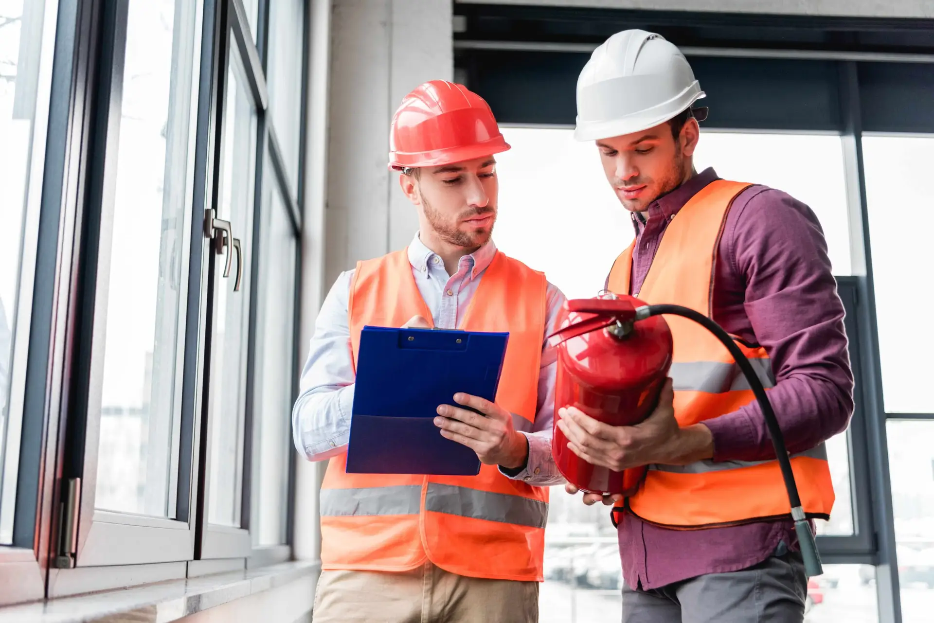 Two safety inspectors wearing hard hats and high‑visibility vests reviewing a clipboard together while examining a fire extinguisher inside a commercial building.