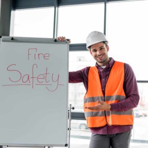A fire safety instructor wearing a hard hat and high‑visibility vest standing beside a whiteboard with “Fire Safety” written on it, gesturing during a training session.