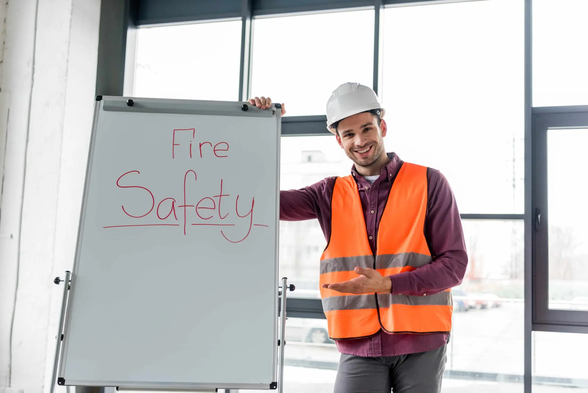A fire safety instructor wearing a hard hat and high‑visibility vest standing beside a whiteboard with “Fire Safety” written on it, gesturing during a training session.