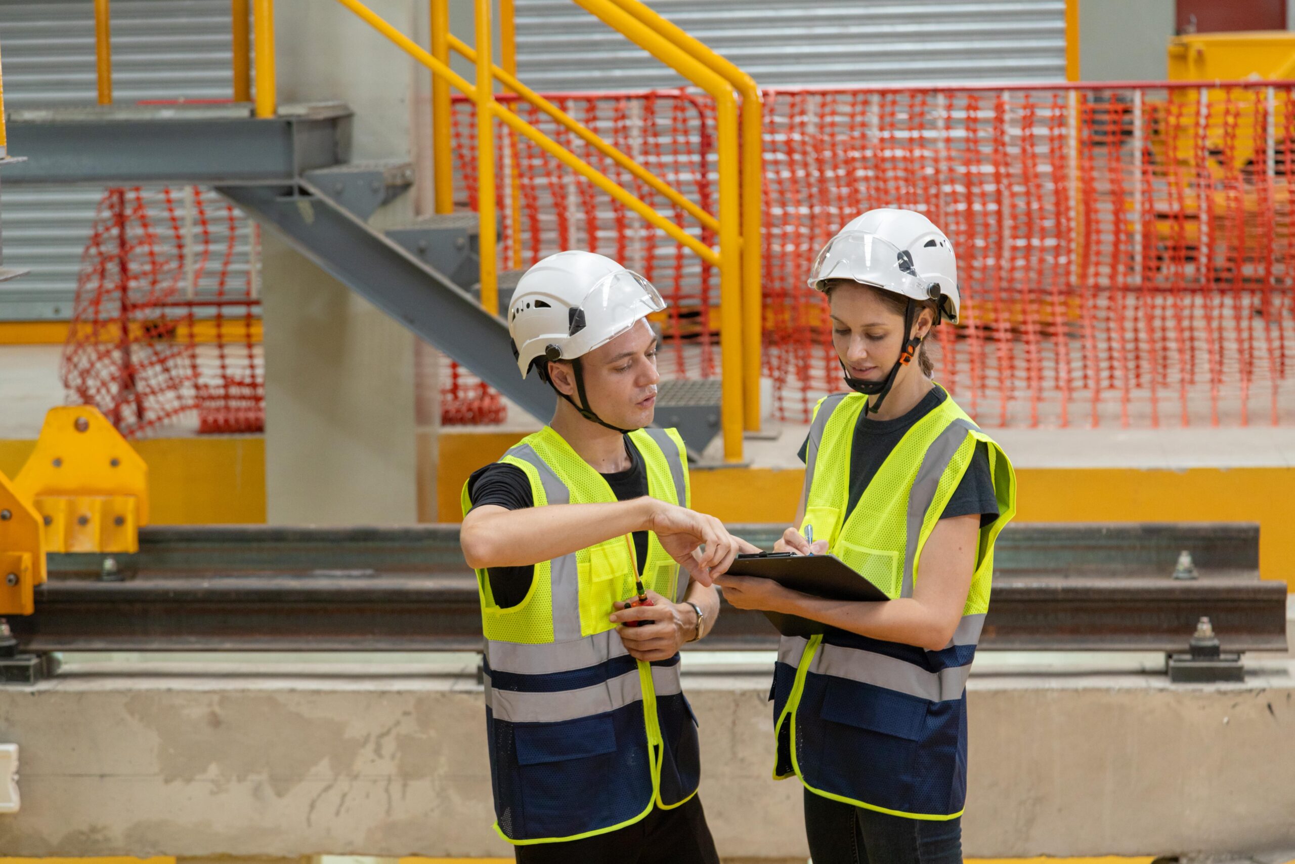 Two construction site workers wearing high-visibility vests and safety helmets reviewing notes on a clipboard in an industrial environment with safety barriers and metal railings.