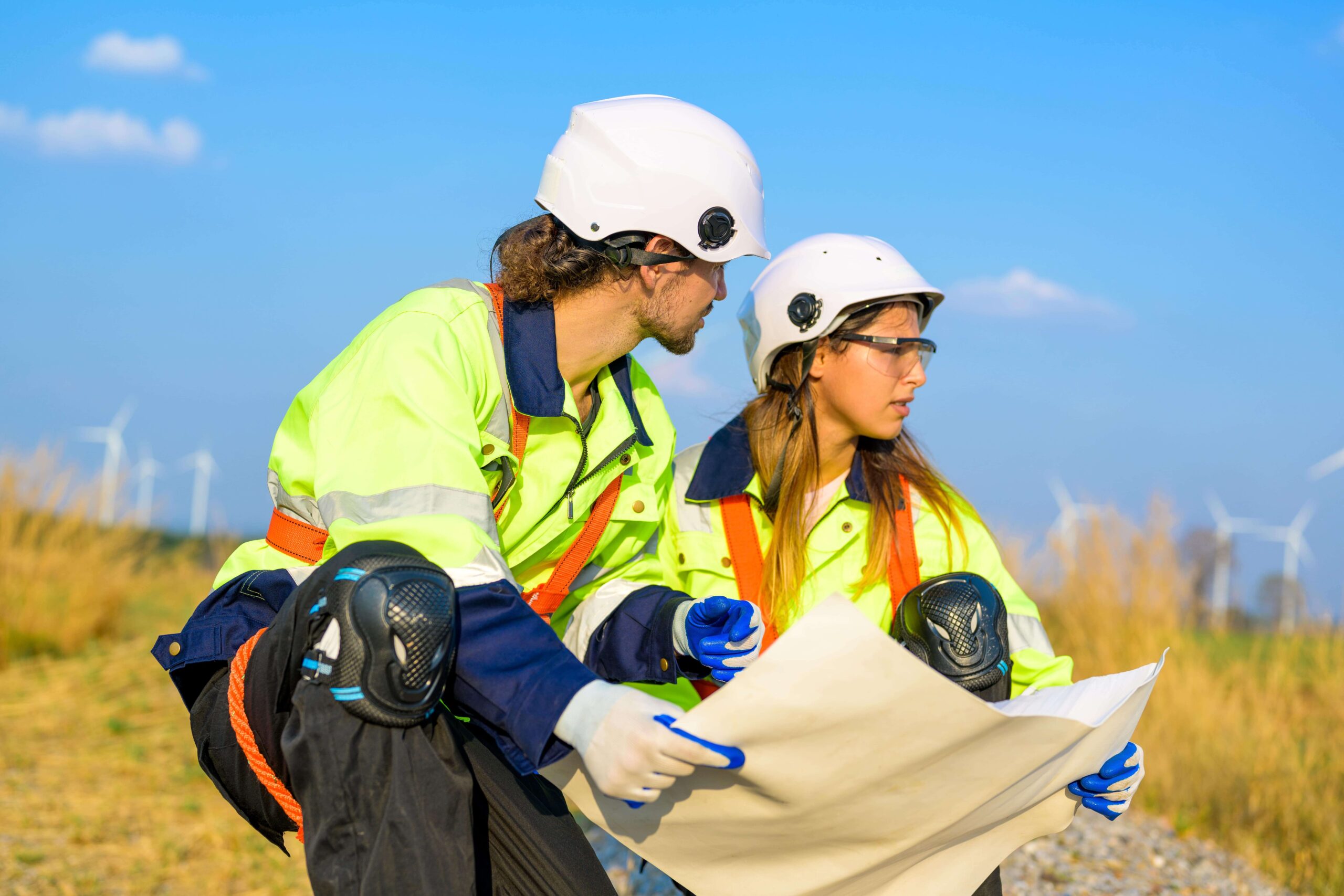 Two environmental safety professionals wearing PPE review site plans in a field with wind turbines in the background.