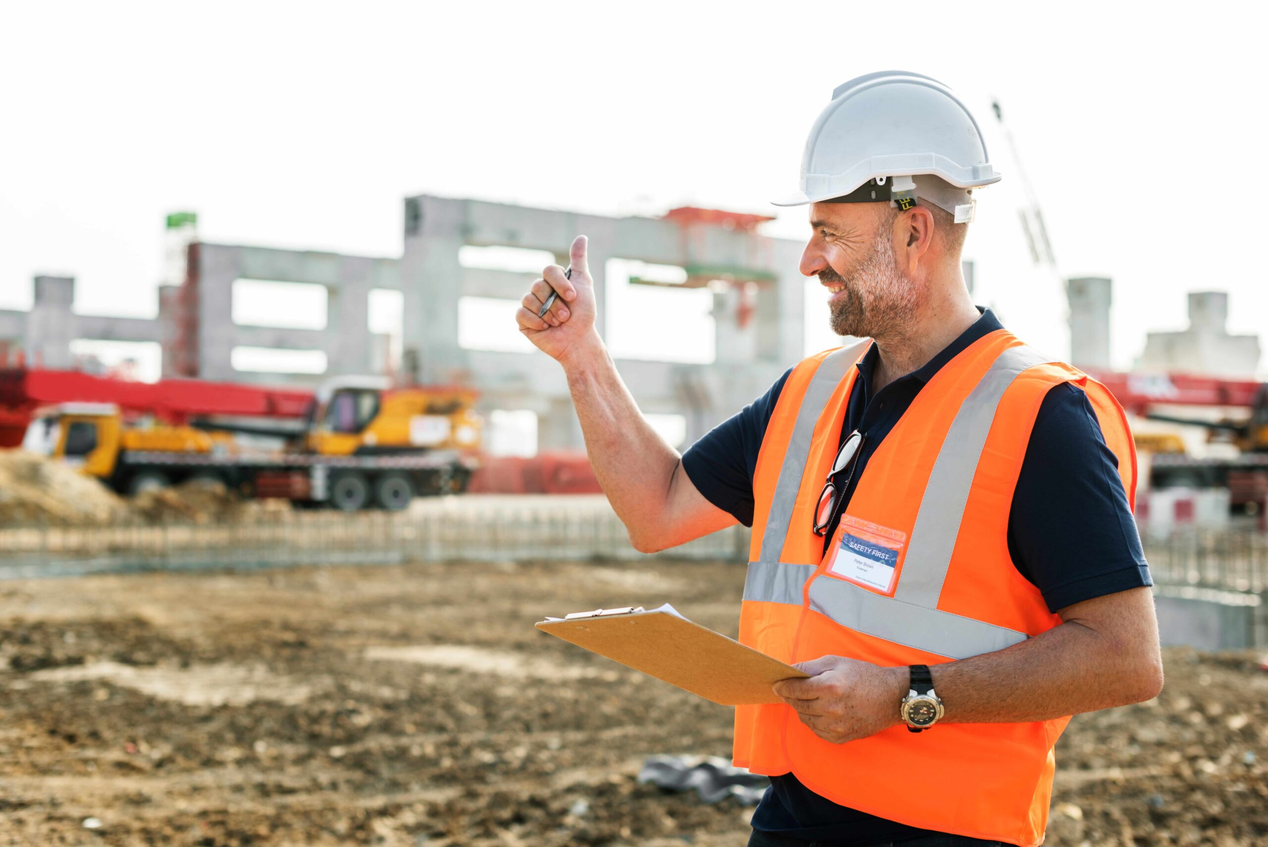 Construction site supervisor in PPE giving a thumbs‑up while holding a clipboard.