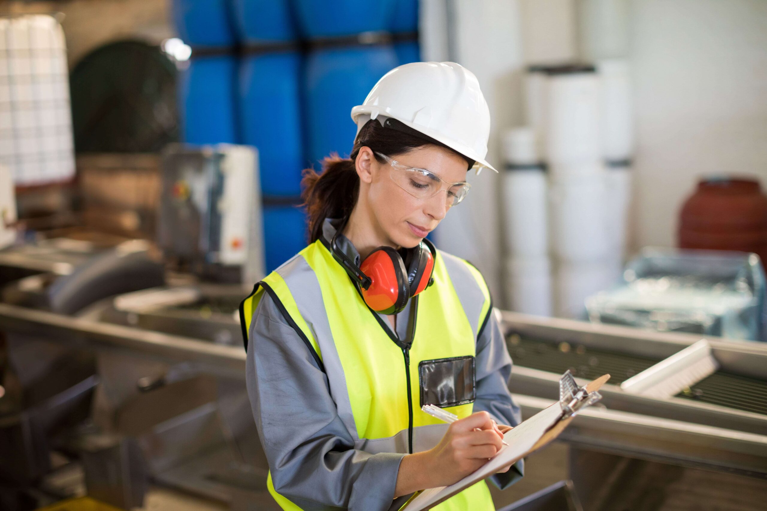 Safety worker in PPE writing notes on a clipboard during an incident investigation in an industrial setting.