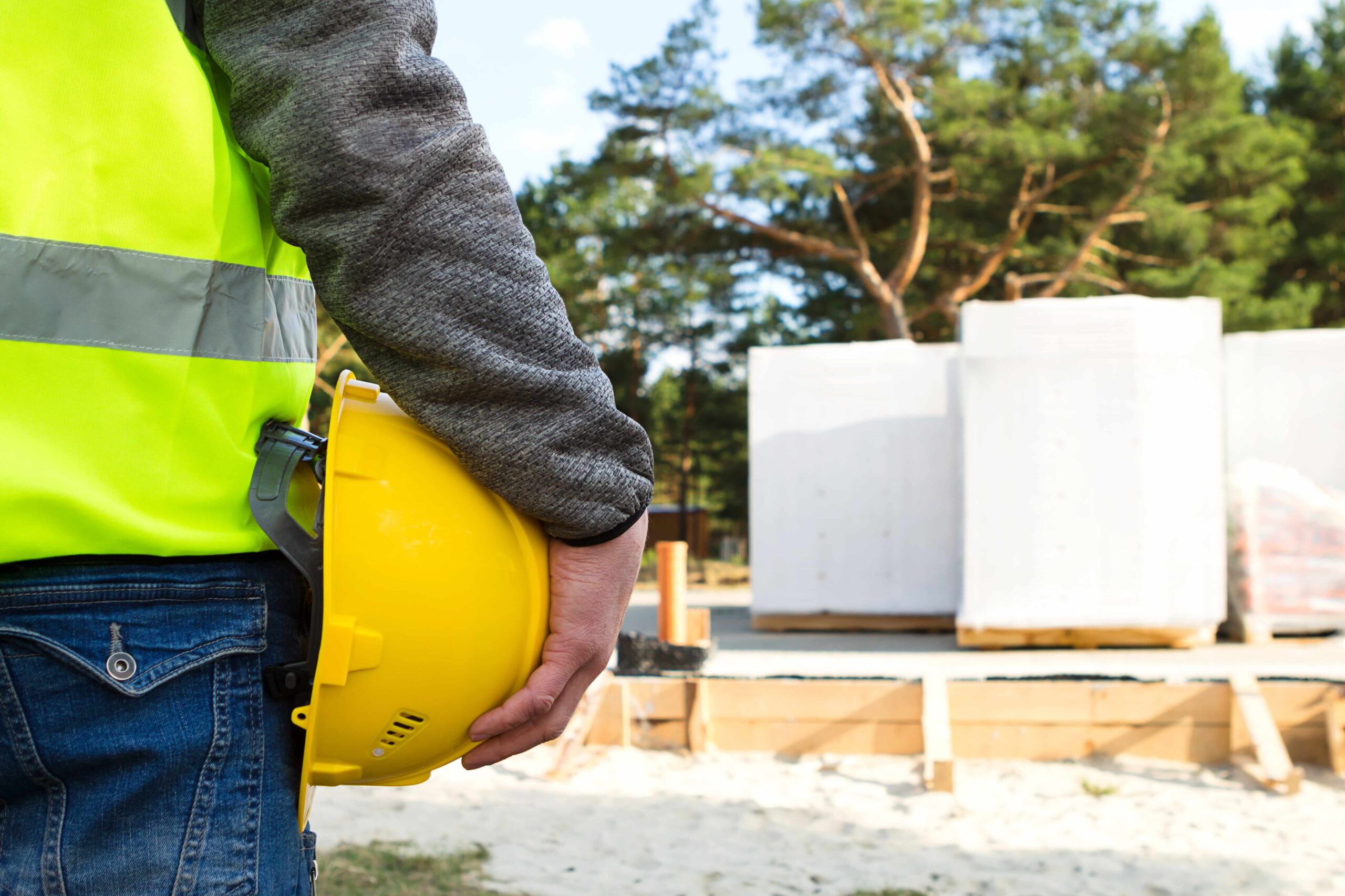 Construction worker holding a yellow hard hat while surveying a worksite with materials in the background.