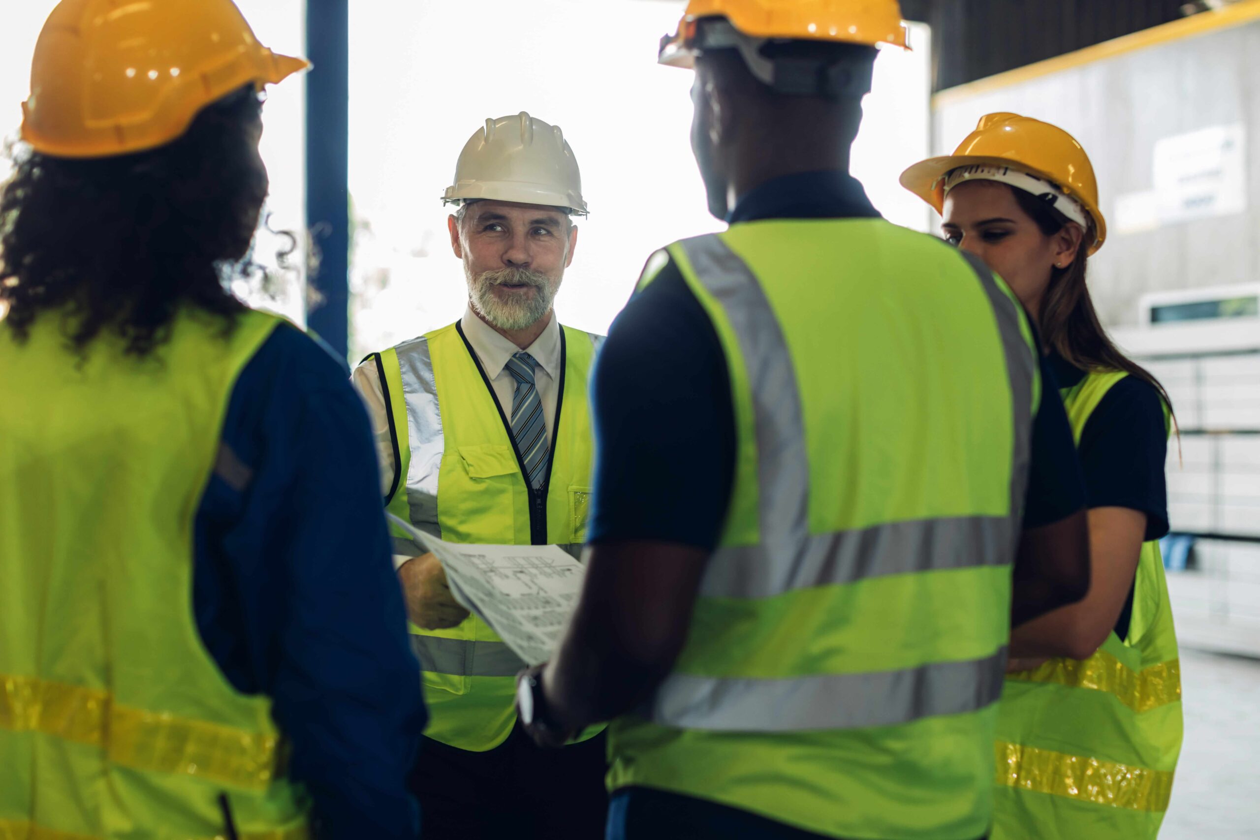 Group of safety professionals in high‑visibility PPE discussing plans with a supervisor at an industrial site.