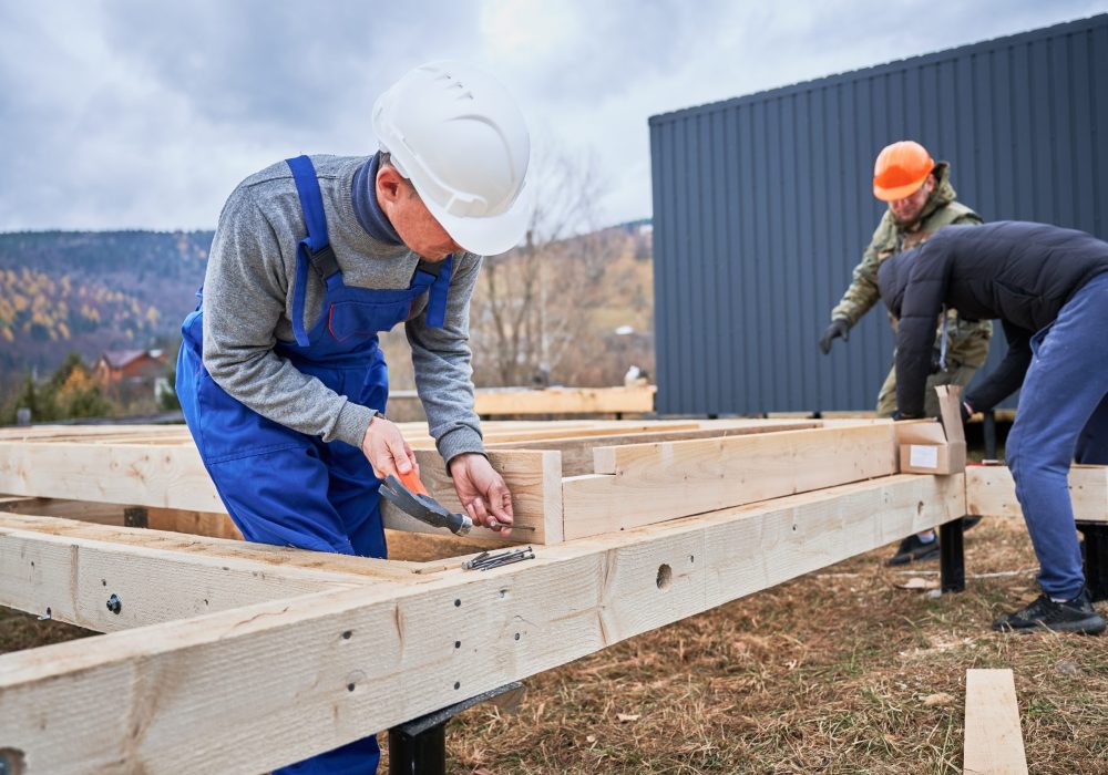 Man worker building wooden frame house on pile foundation. Carpenter hammering nail into wooden plank, using hammer. Carpentry concept.