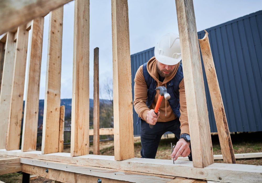 The image shows a construction worker wearing a white hard hat and a brown hoodie, using a hammer to drive a nail into wooden beams. The worker is building or assembling a wooden structure, with vertical wooden planks and horizontal supports visible. In the background, there is a blue container or building.