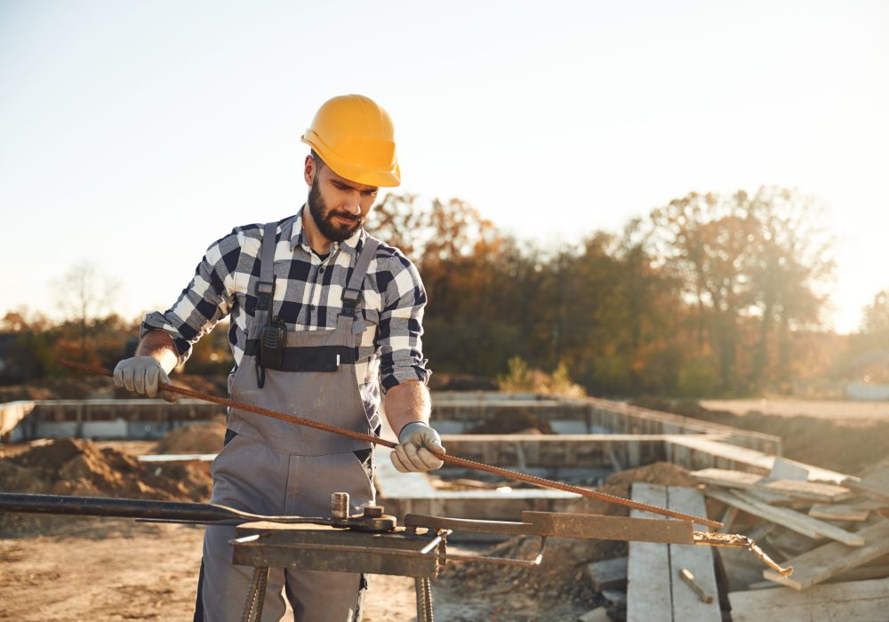 A construction worker wearing a yellow hard hat, checkered shirt, and gray overalls is bending a metal rod using a manual rebar bender at a construction site. The background shows an unfinished building foundation and scattered construction materials with trees in the distance under bright sunlight.